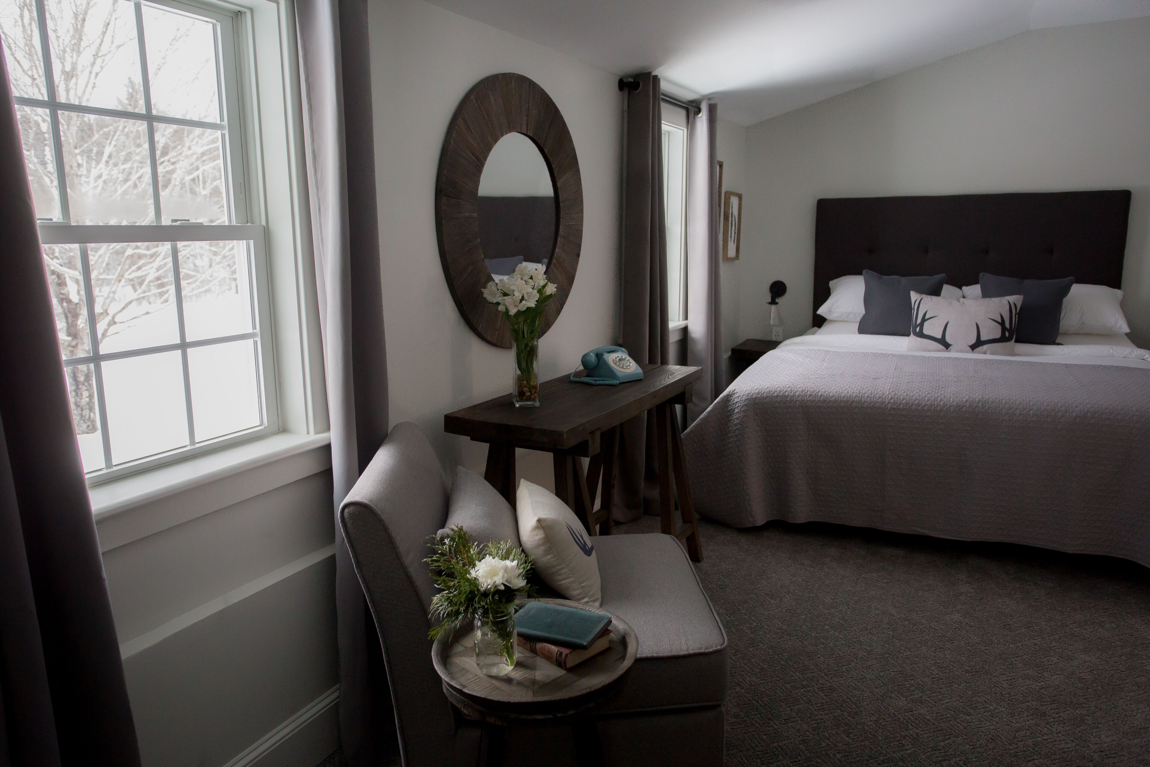 A bedroom with a large bed, a bedside table with a vase of flowers, a chair with cushions, and a window with curtains.