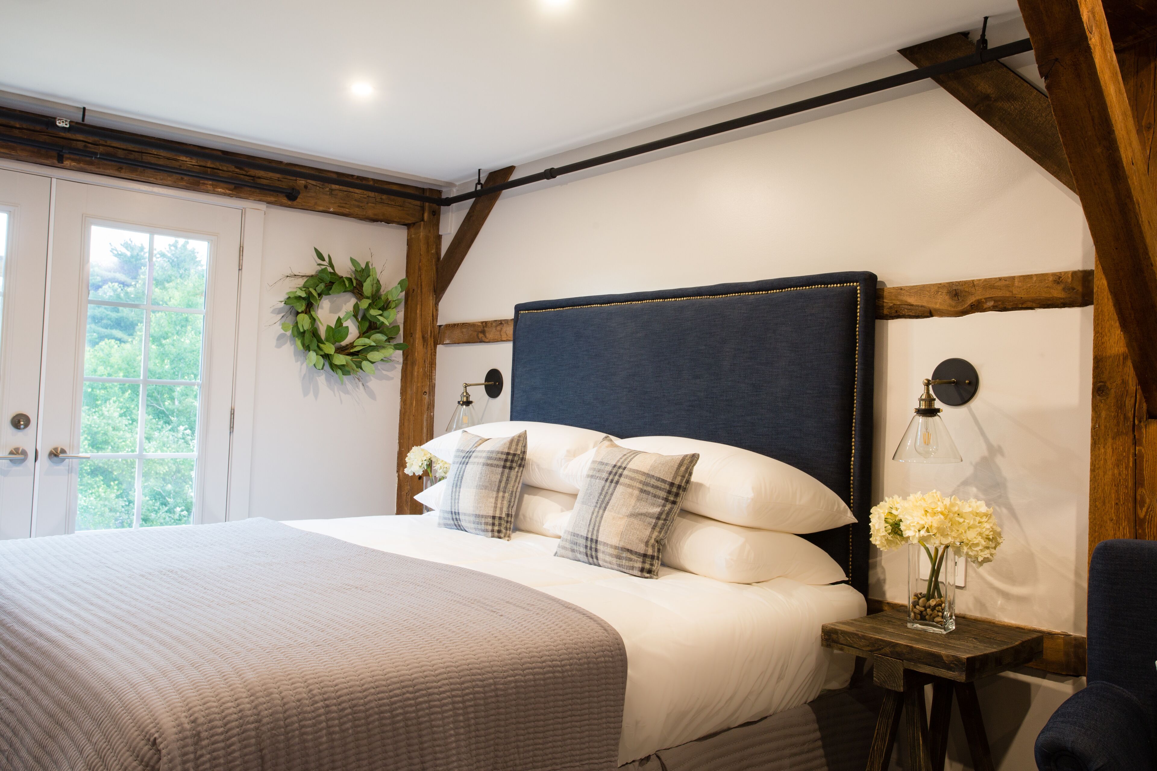 A bedroom with a large bed, a wooden headboard, a bedside table with a lamp, and a window with a view of greenery.