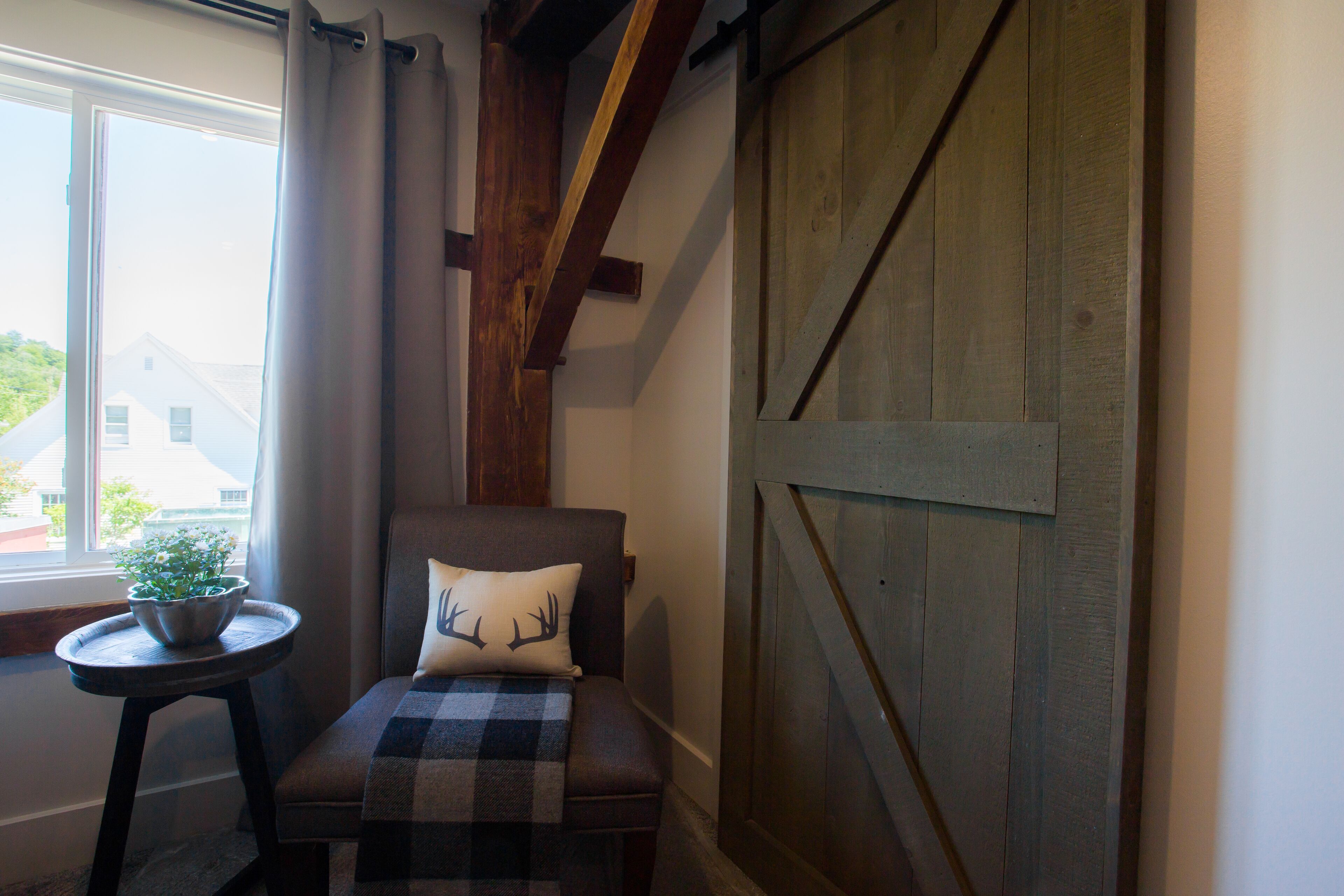 Una habitación acogedora con ventana de madera, una mesita redonda con una planta, una manta escocesa y un cojín con un diseño de ciervo.
