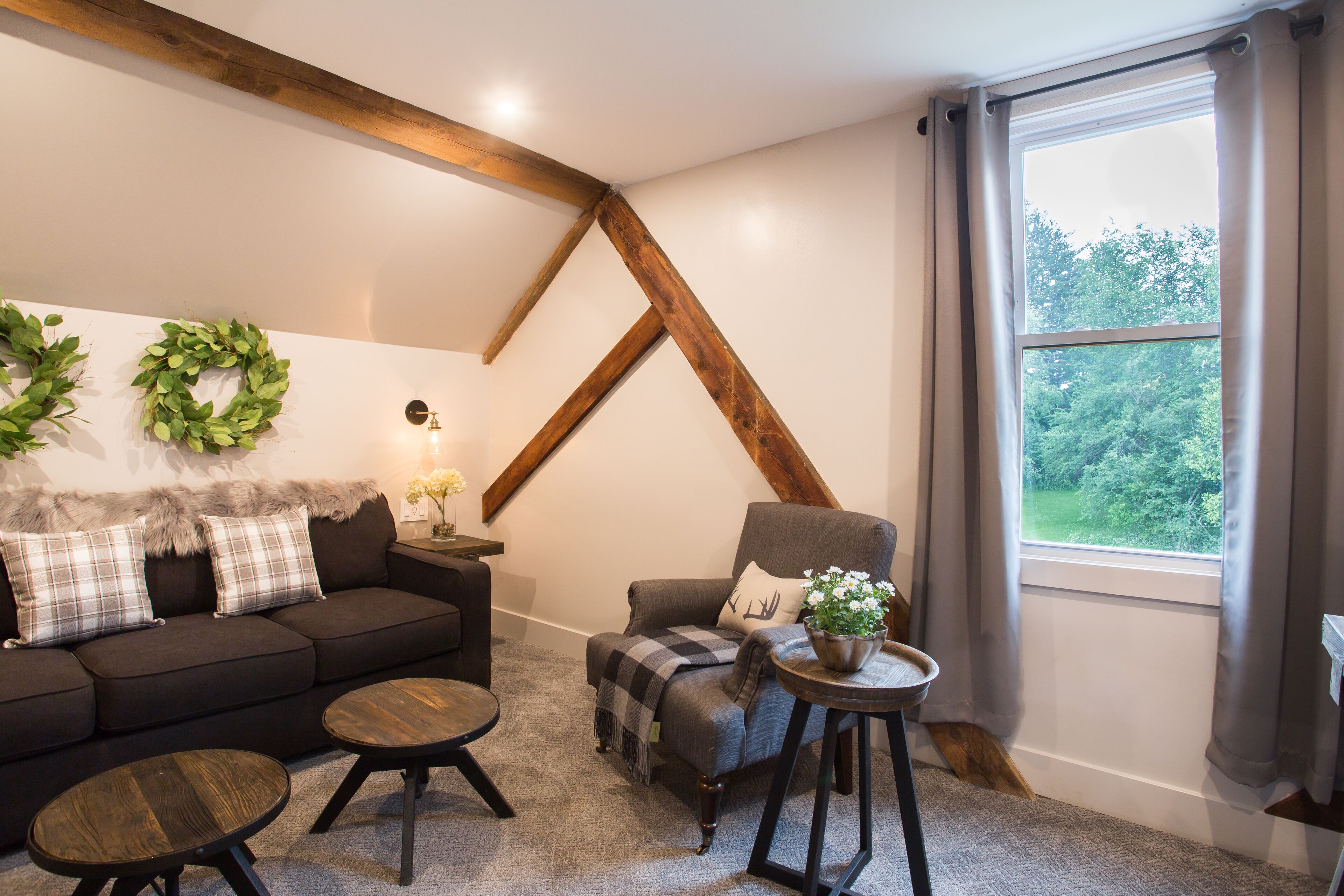 Un salon chaleureux avec un canapé foncé, deux tables rondes, un fauteuil gris et une grande fenêtre avec des rideaux.