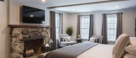 A bedroom with a stone fireplace, a large bed, a TV mounted above the fireplace, and a window with curtains.