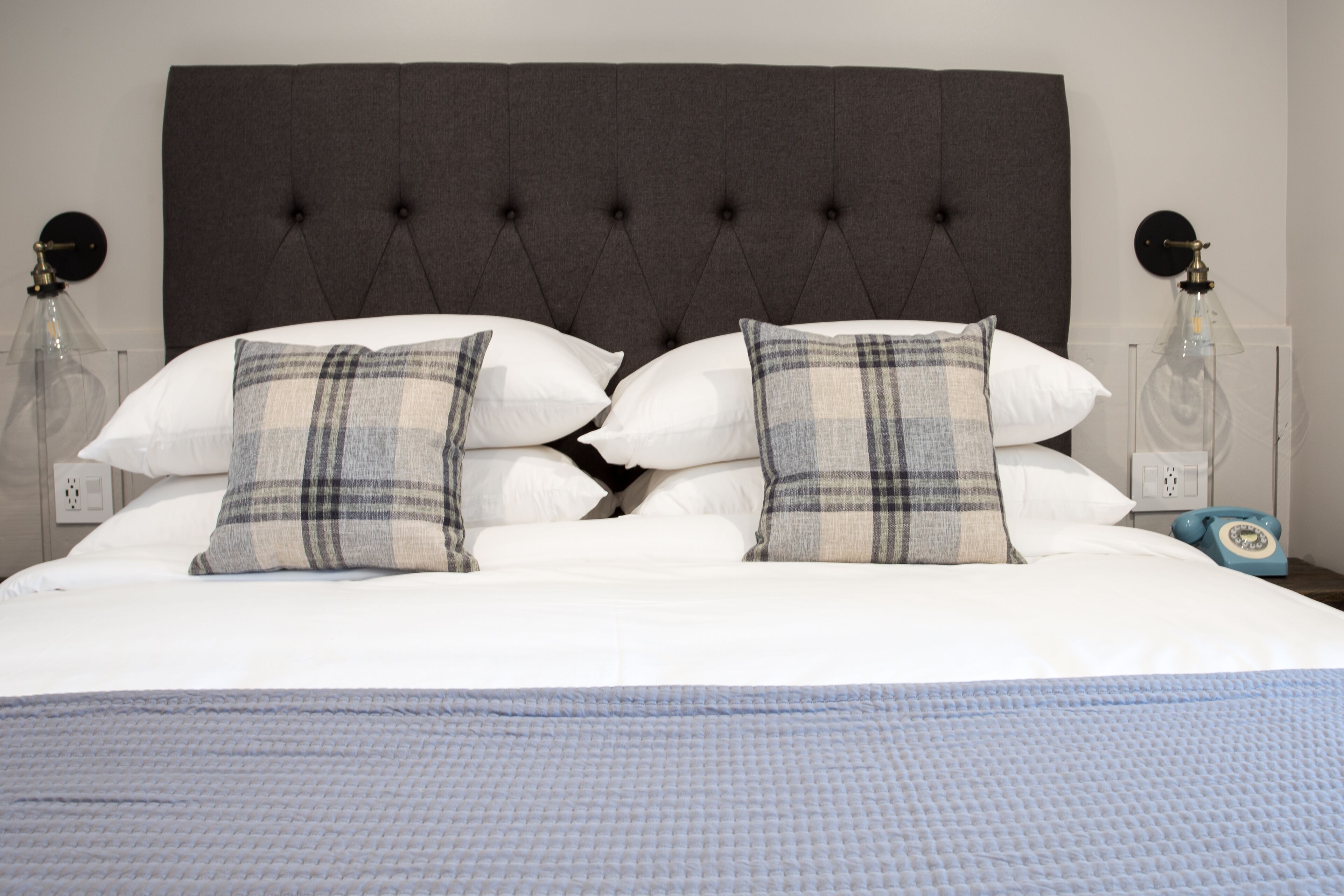 A neatly made bed with a tufted headboard, white and plaid pillows, and a blue textured blanket.