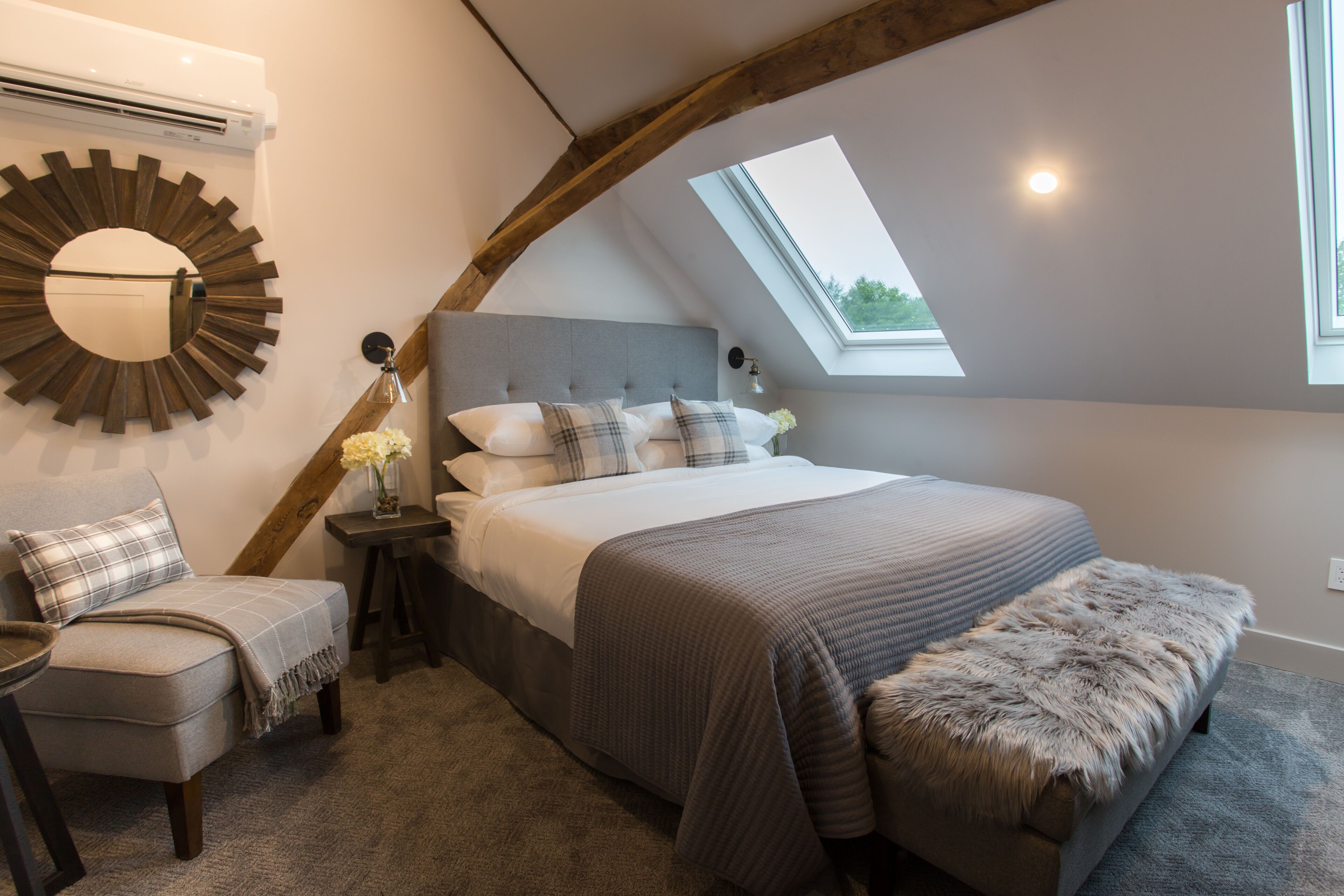 A bedroom with a skylight, a bed with a grey headboard, a chair, a small table, and a decorative mirror.