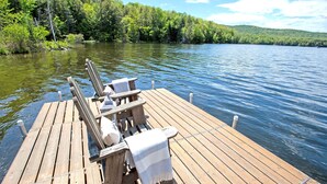 A wooden dock with two Adirondack chairs and a blanket, overlooking a calm lake with dense forest in the background.