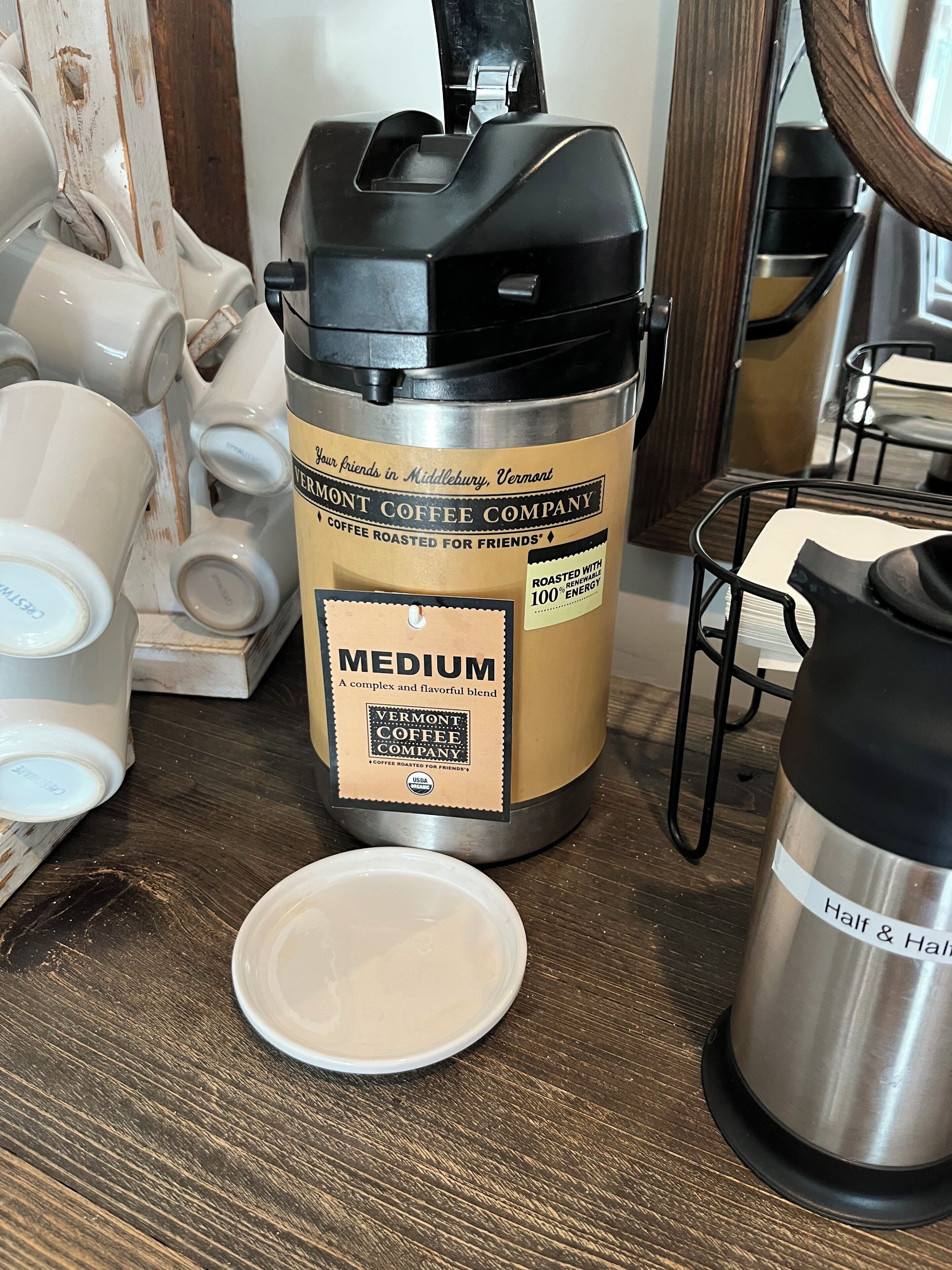 Une machine à café portant une étiquette indiquant qu’elle est de la marque Vermont Coffee Company, un thermos en acier inoxydable et une assiette blanche posée sur une table en bois.