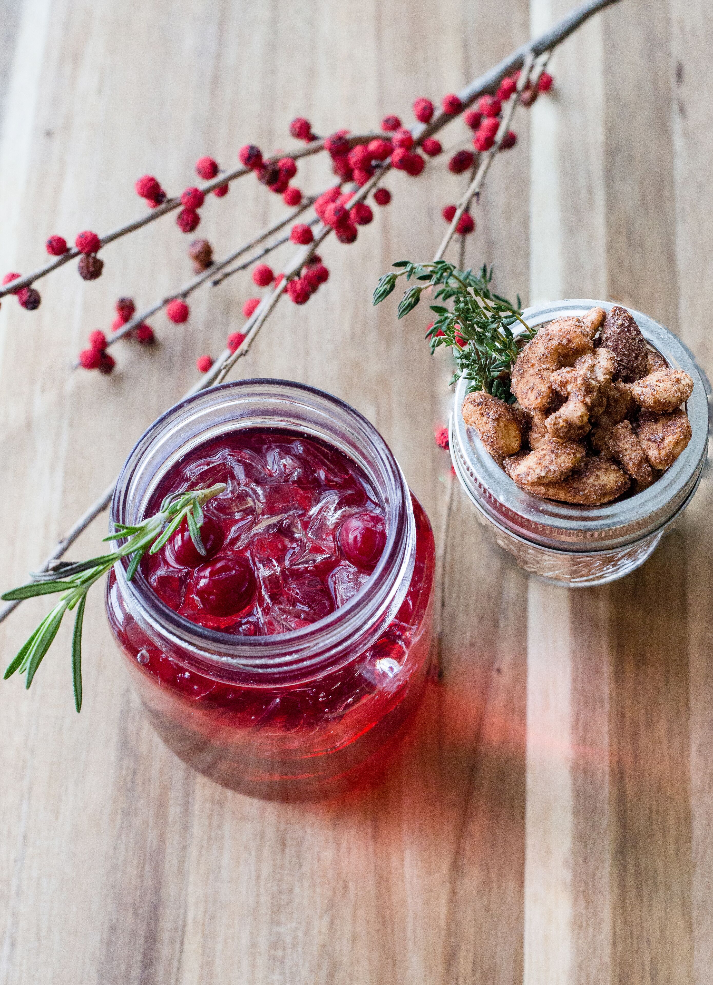 A jar of red fruit preserve with a sprig of rosemary, a bowl of breaded chicken, and a branch with red berries on a wooden surface.