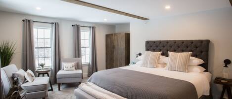 A bedroom with a large bed, a bench, a chair, a wardrobe, and a small table with a lamp.