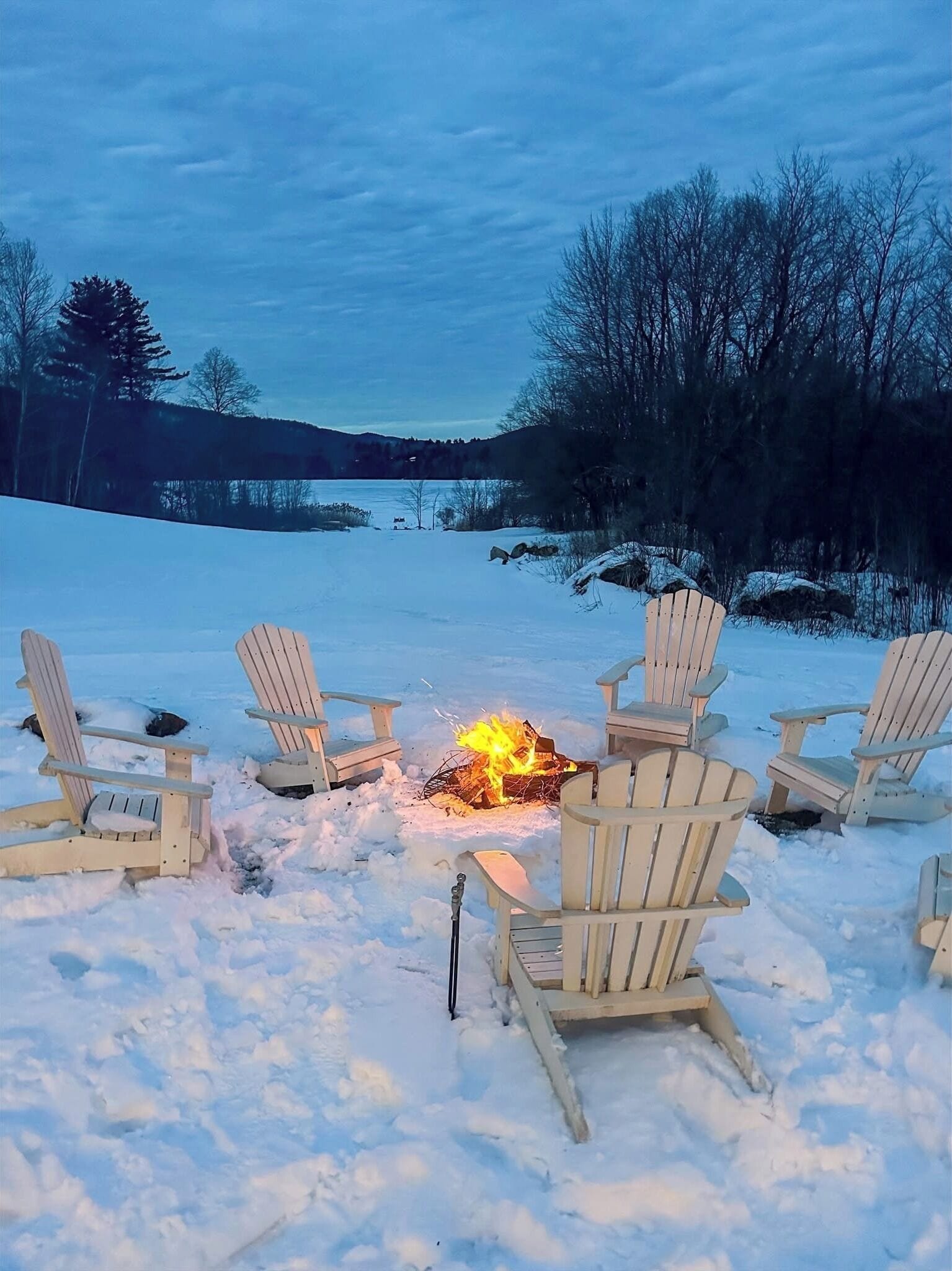 A snow-covered outdoor area with wooden chairs arranged around a campfire.