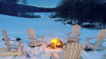 A snow-covered outdoor area with wooden chairs arranged around a campfire.