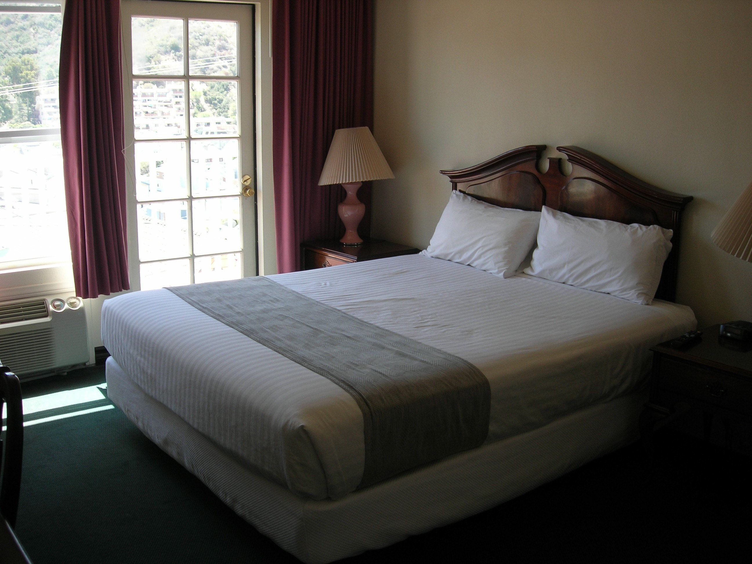 A neatly made bed with a wooden headboard, a bedside lamp, and a view of a building through a window with curtains.