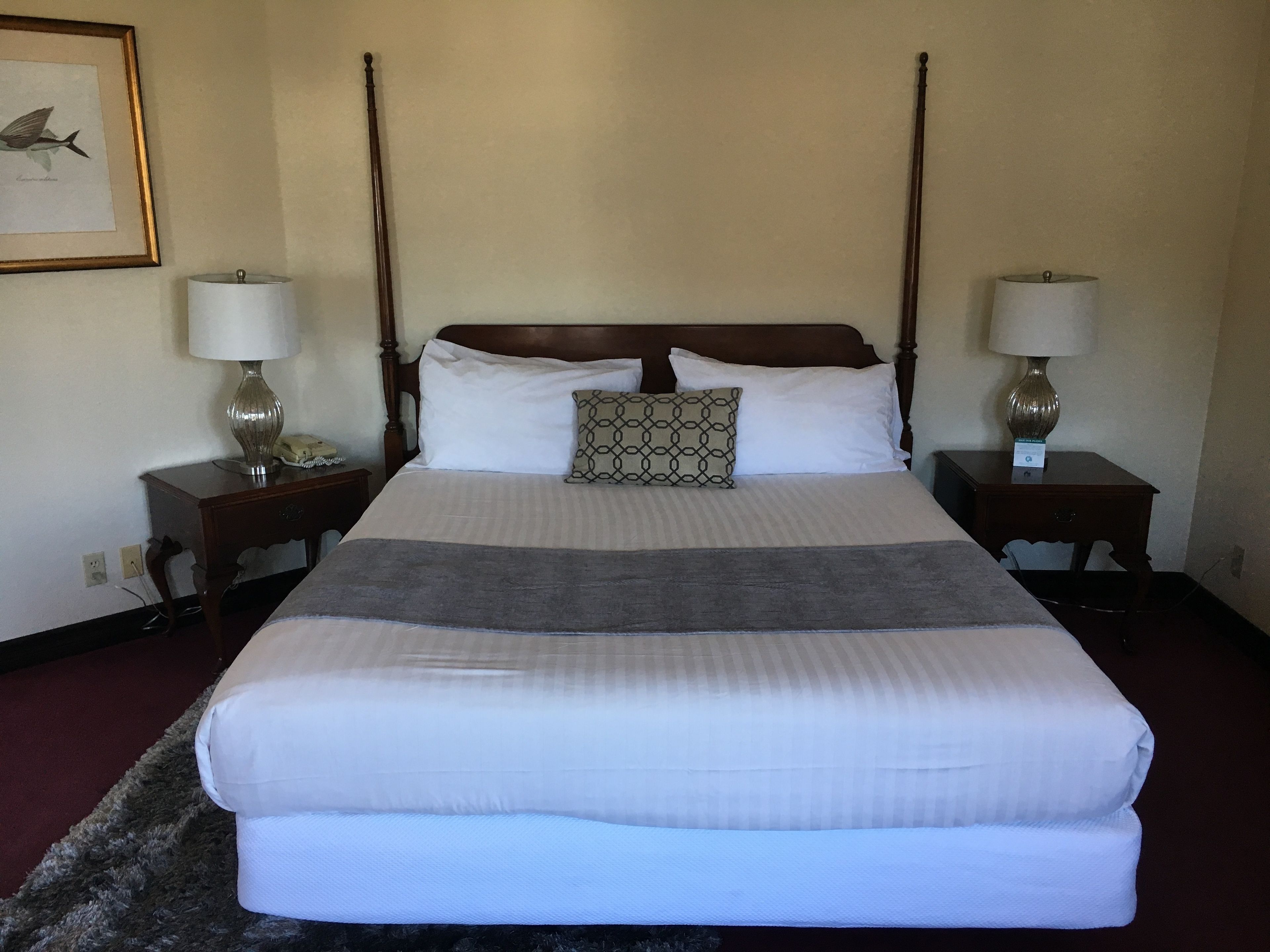 A neatly made bed with a headboard, two bedside tables with lamps, a patterned pillow, and a framed picture on the wall.
