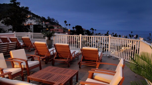 A terrace with wooden furniture, a white railing, and a view of the sea and buildings in the background. - Hotel St. Lauren (Avalon)