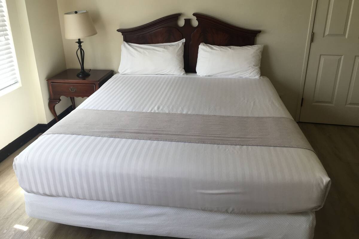 Una cama bien hecha con cabecera de madera, una mesita de noche con lámpara, y una puerta al fondo.