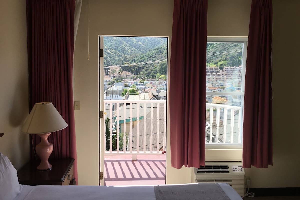 Habitación de hotel con balcón, cama, mesita de noche con lámpara y vista a un paisaje urbano y montañas.