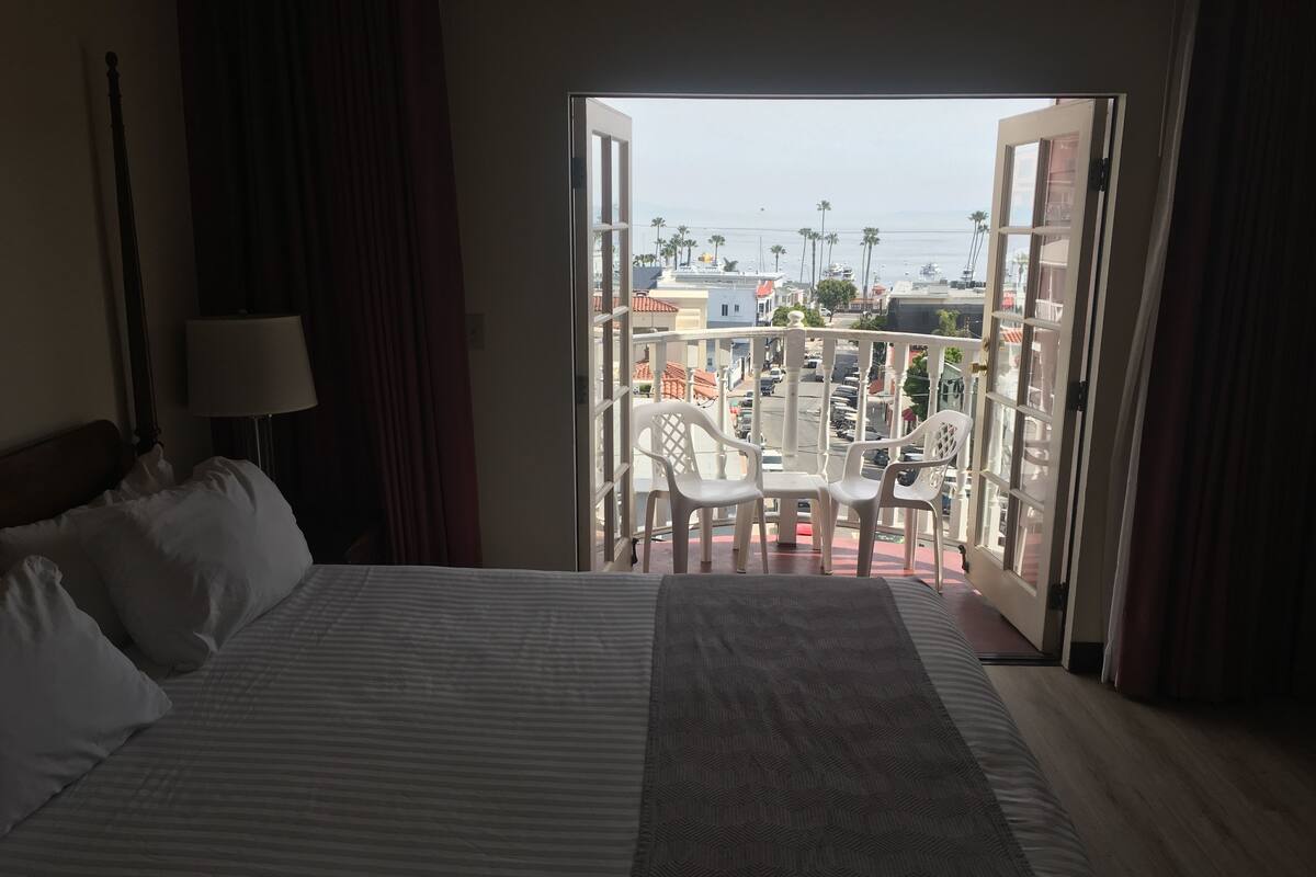 Habitación de hotel con cama, mesita de noche, lámpara y vistas a un balcón con sillas y una mesa.