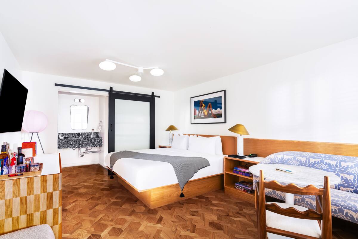 A hotel room with a bed, a desk, a chair, a bookshelf, a mirror, a bathroom, and a painting on the wall.