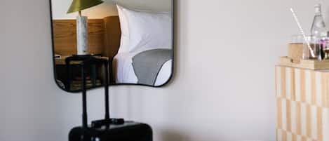 Quarto de hotel com uma cama, mala, um banquinho quadriculado e um espelho refletindo o interior do quarto.