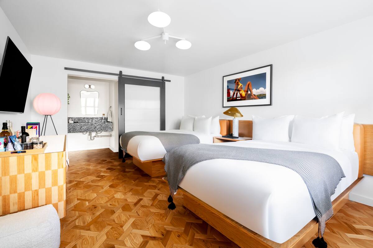A modern hotel room with two beds, a wooden floor, a desk with toiletries, a TV, and a painting on the wall.