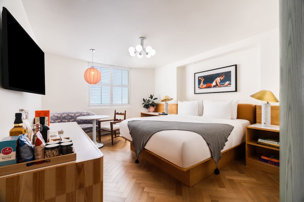 A modern hotel room with a large bed, bedside tables, a desk, and a chair. There is a window with blinds, a ceiling light, and a painting on the wall.