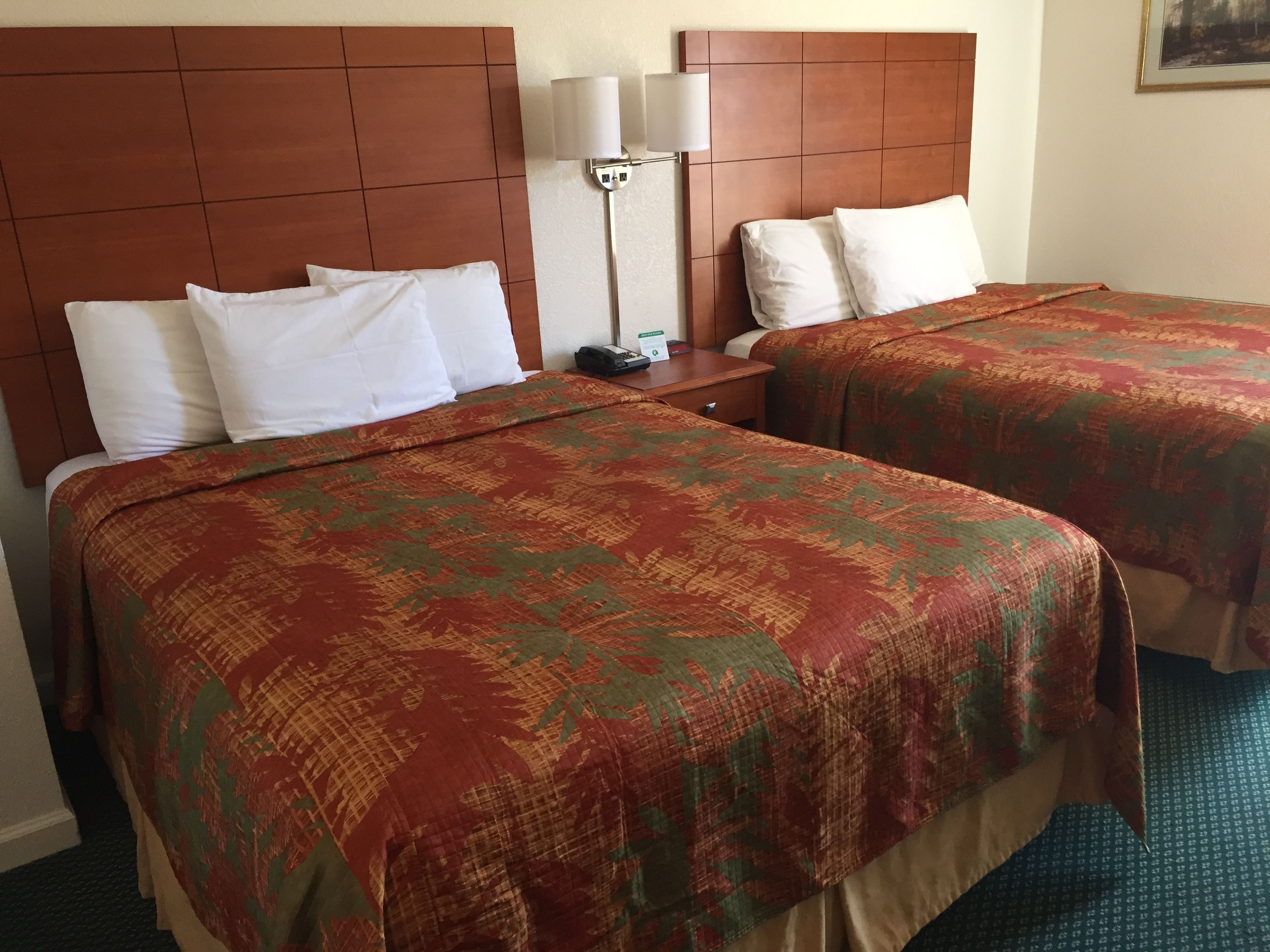 A hotel room with two beds, each with white pillows and a patterned bedspread. There is a nightstand with a lamp and a sign indicating the room number.