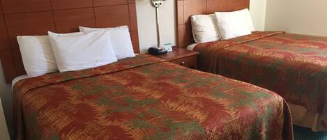 A hotel room with two beds, each with white pillows and a patterned bedspread. There is a nightstand with a lamp and a sign indicating the room number.