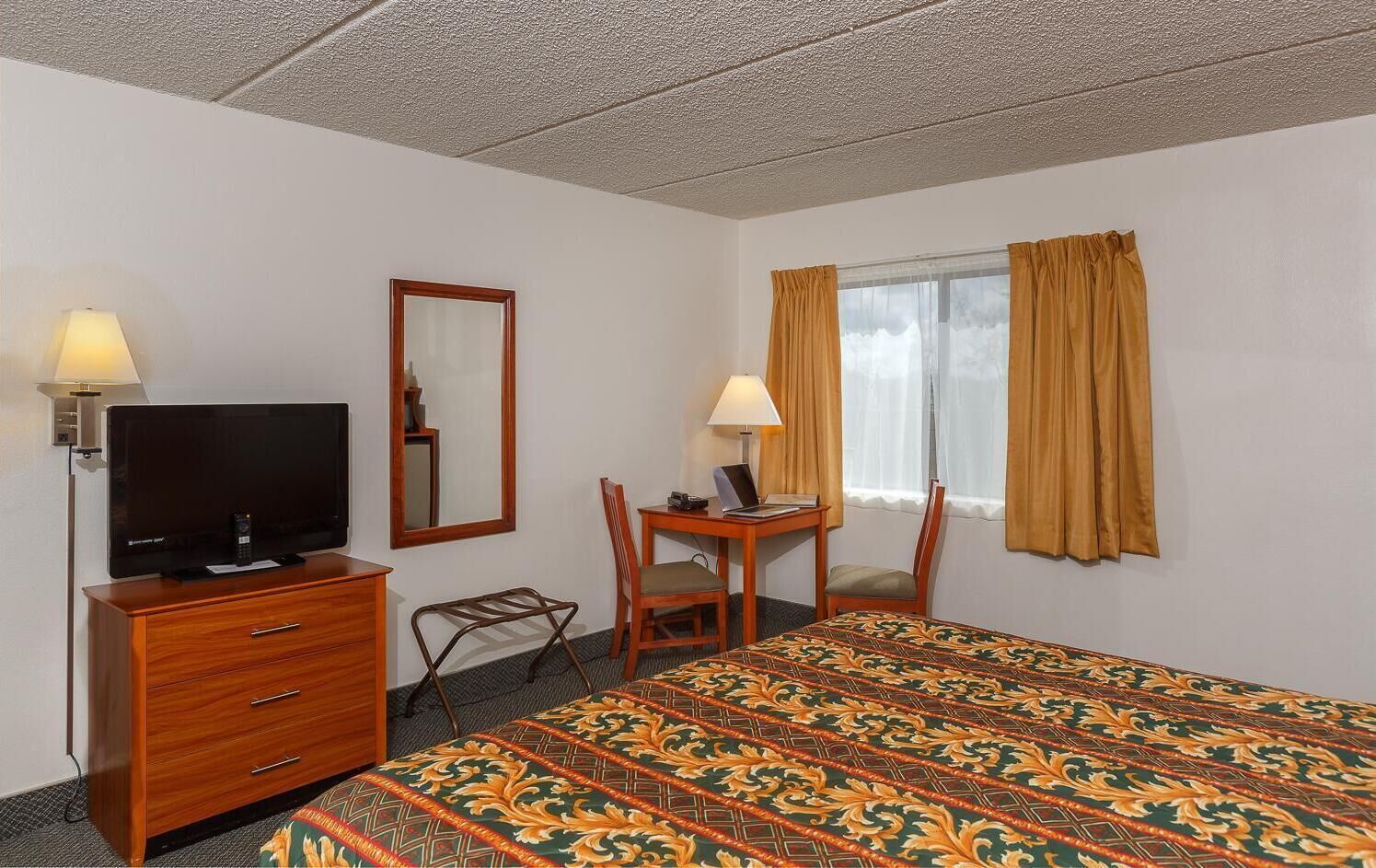 A hotel room with a bed, a TV, a desk with a chair, a lamp, and a window with curtains.