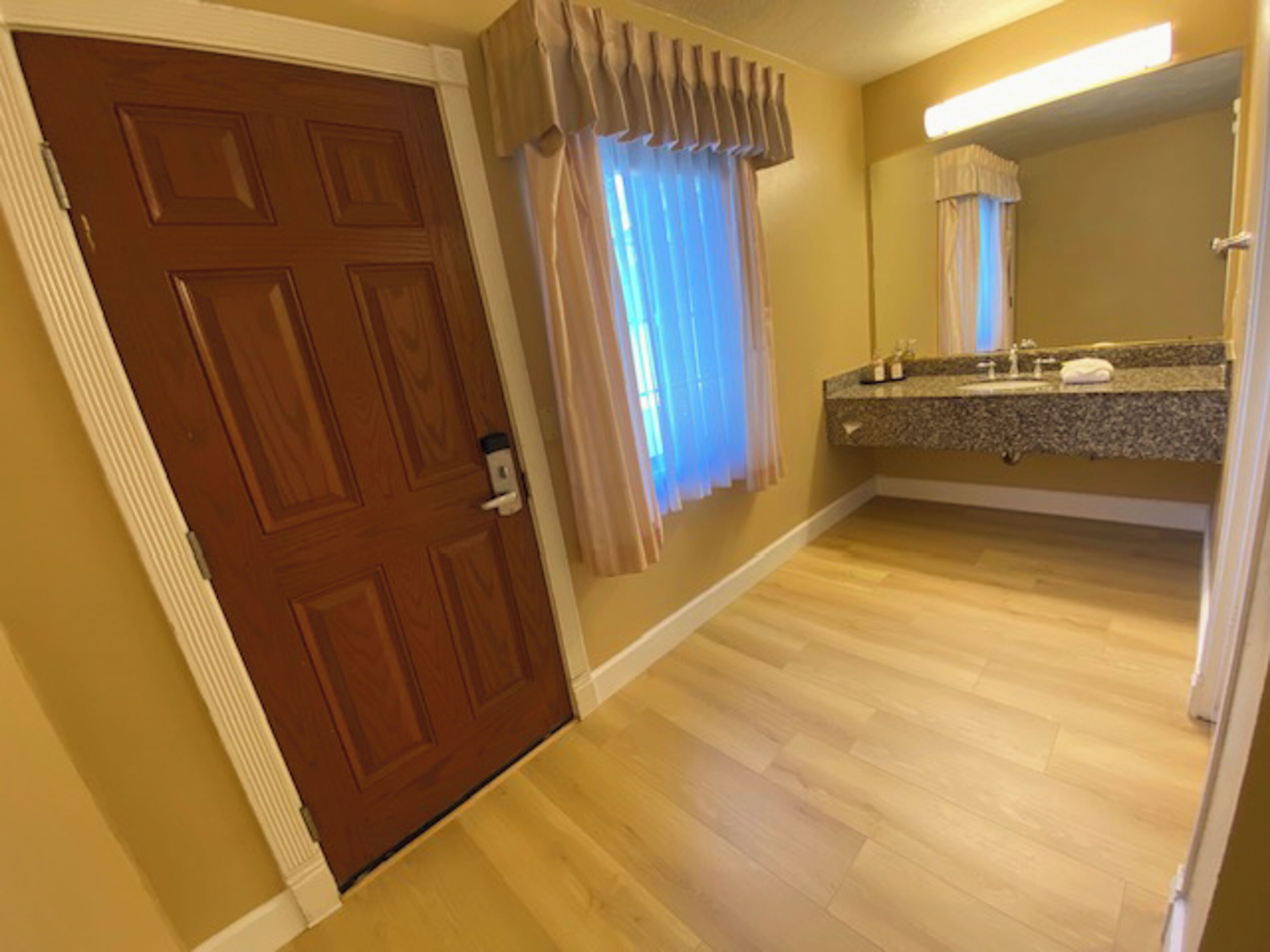 Una habitación de hotel con puerta de madera, baño con tocador y espejo, y ventana con cortinas.