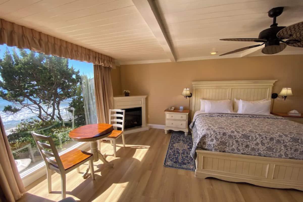 A bedroom with a bed, bedside tables, a chair, a table, a ceiling fan, and a view of the outdoors.