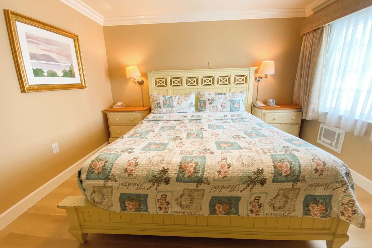 A bedroom with a bed, two bedside tables, a nightstand, a wall-mounted lamp, a framed picture, and a window with curtains.