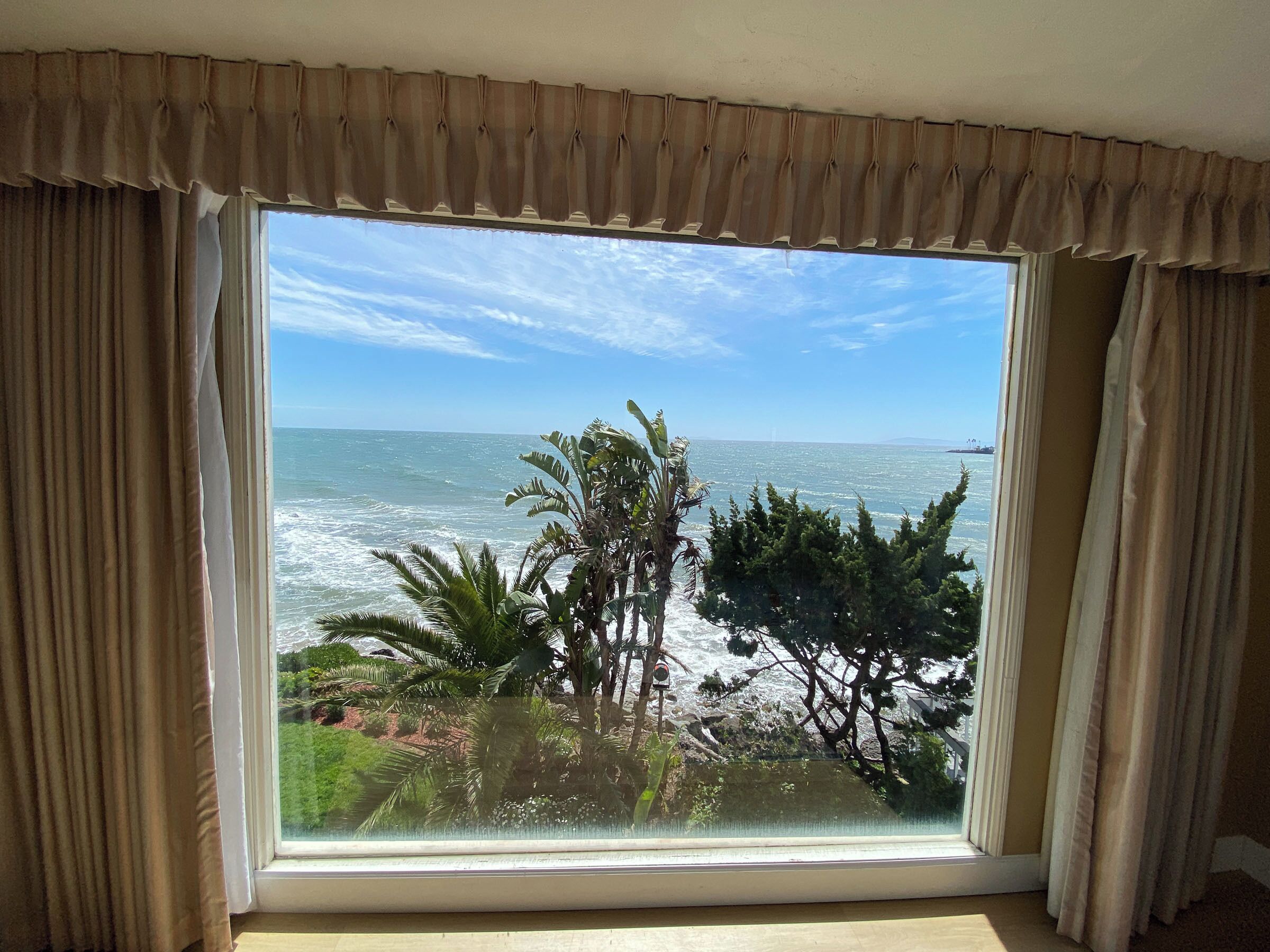 Una habitación con un ventanal grande que da al océano, con cortinas y vistas a palmeras y follaje.