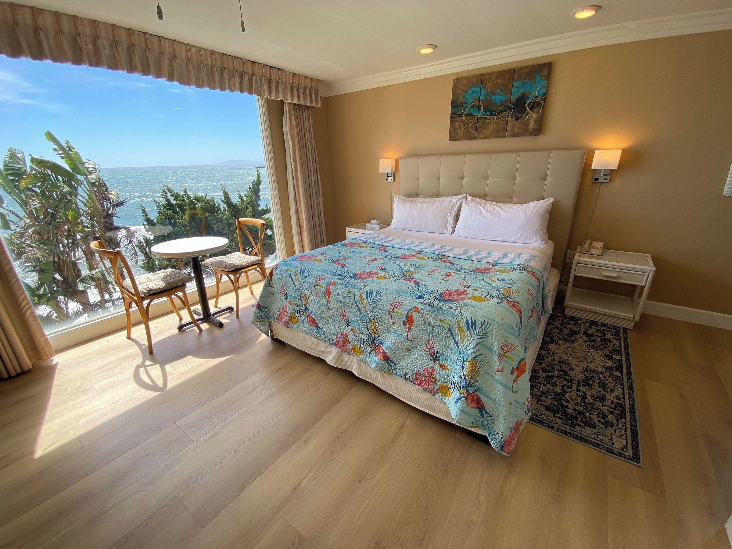 A bedroom with a large bed, a view of the ocean, a dining table with chairs, and a small rug.