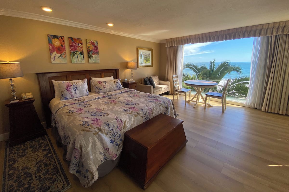 A bedroom with a large bed, a wooden bench, a small table, and a view of the ocean.