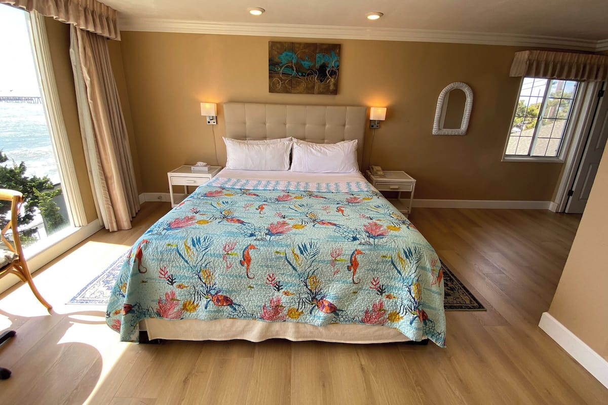A bedroom with a large bed, two bedside tables, a headboard, a painting, and a view of the outdoors.