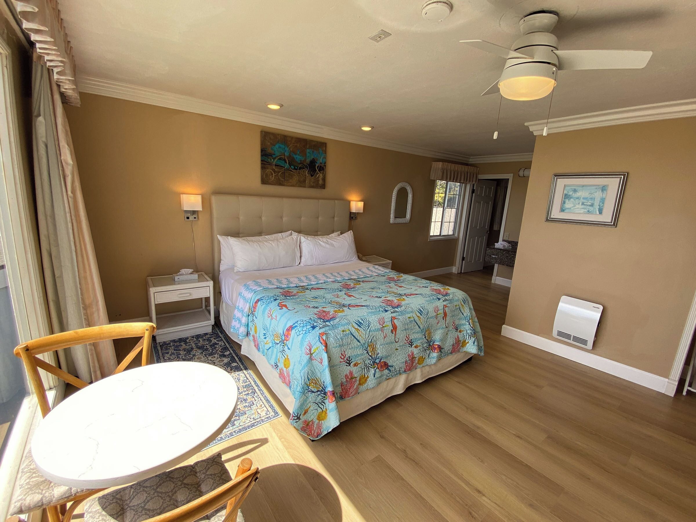 A hotel room with a bed, a round table, a chair, a ceiling fan, and a painting on the wall.