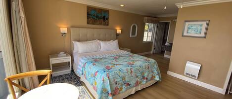 A hotel room with a bed, a round table, a chair, a ceiling fan, and a painting on the wall.