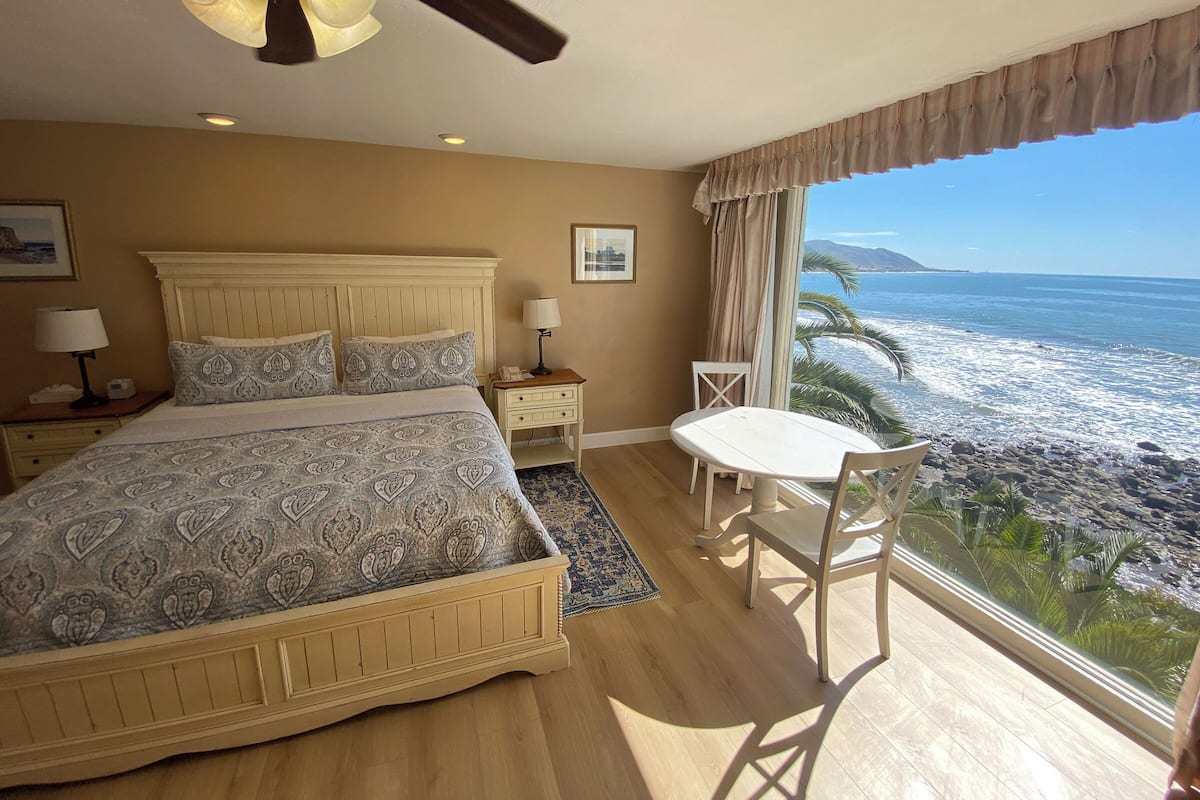 A bedroom with a bed, bedside tables, a nightstand, a window with a view of the ocean, and a ceiling fan.