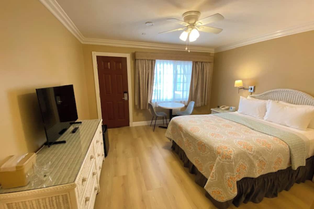 A hotel room with a bed, a desk with a television, a chair, a table, and a ceiling fan.