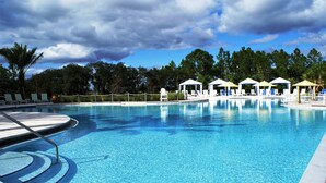 A clear swimming pool with lounge chairs and umbrellas, surrounded by trees and a cloudy sky.
