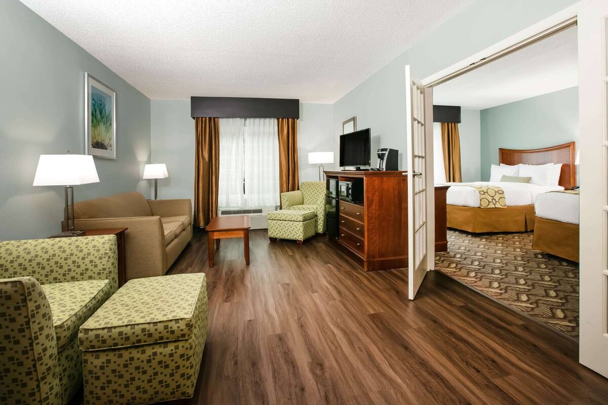 Une chambre d’hôtel avec un canapé, un fauteuil, une petite table, une télévision et un lit recouvert d’un tapis à motifs.