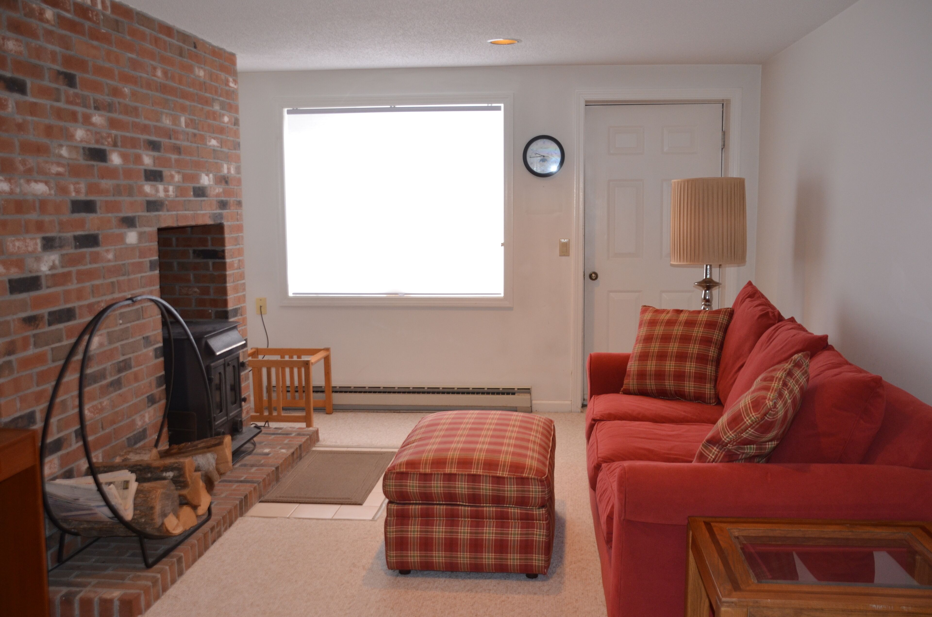 A living room with a brick fireplace, a red couch, a plaid ottoman, and a window with blinds.