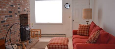 A living room with a brick fireplace, a red couch, a plaid ottoman, and a window with blinds.