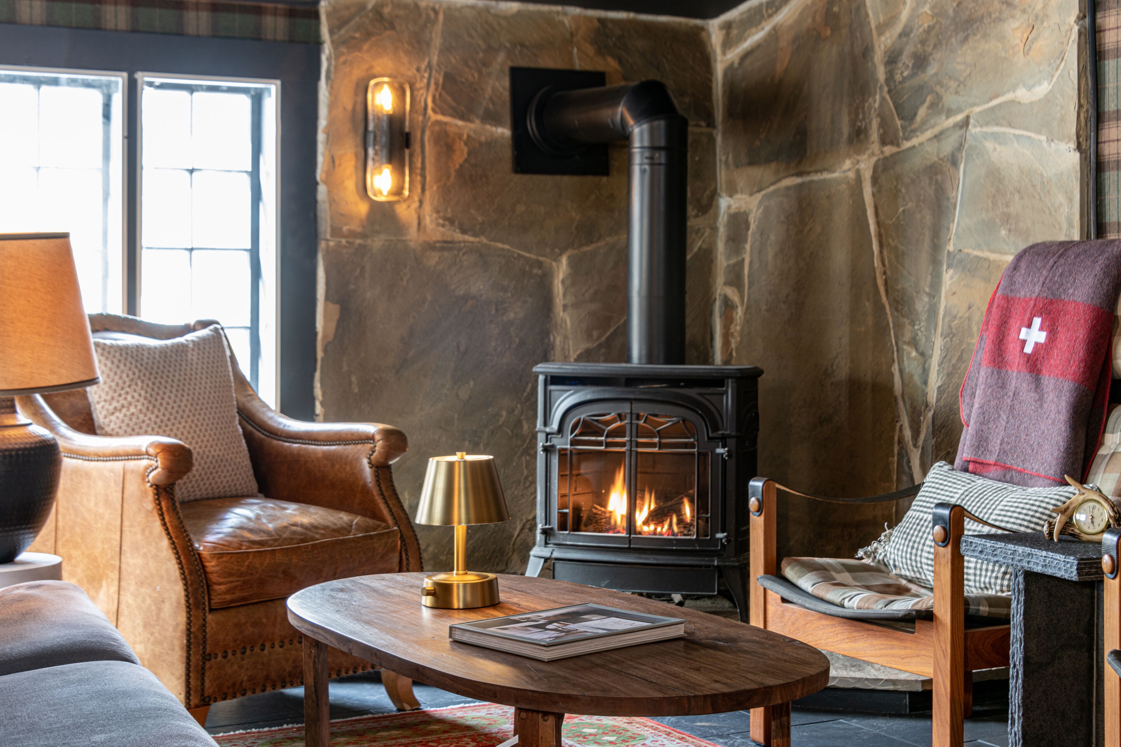 Ein gemütlicher Raum mit Steinwand, Kamin, Ledersesseln, einem Holztisch und einer Lampe.