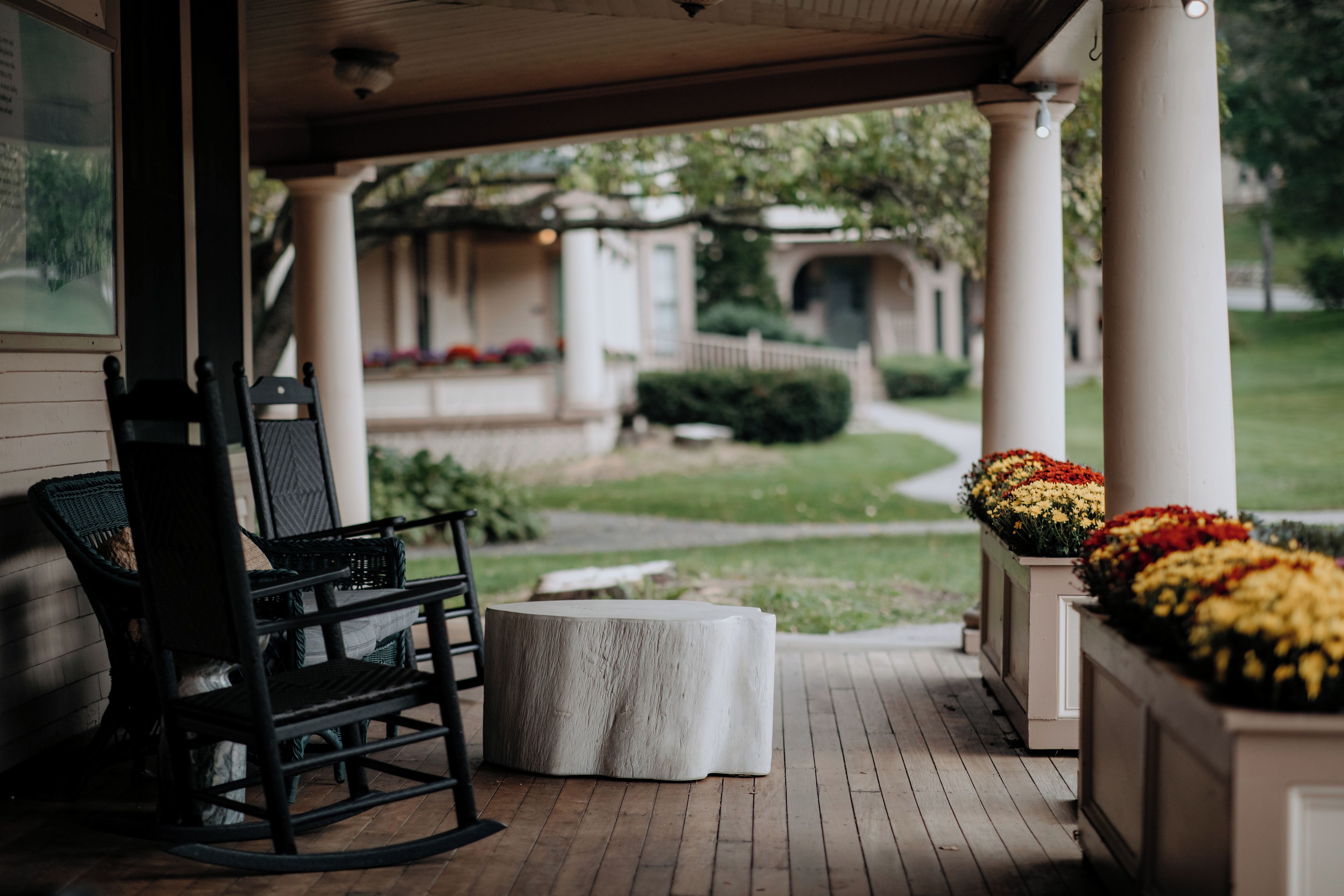 A covered porch with a rocking chair and a round table, overlooking a well-maintained lawn and a building in the background.