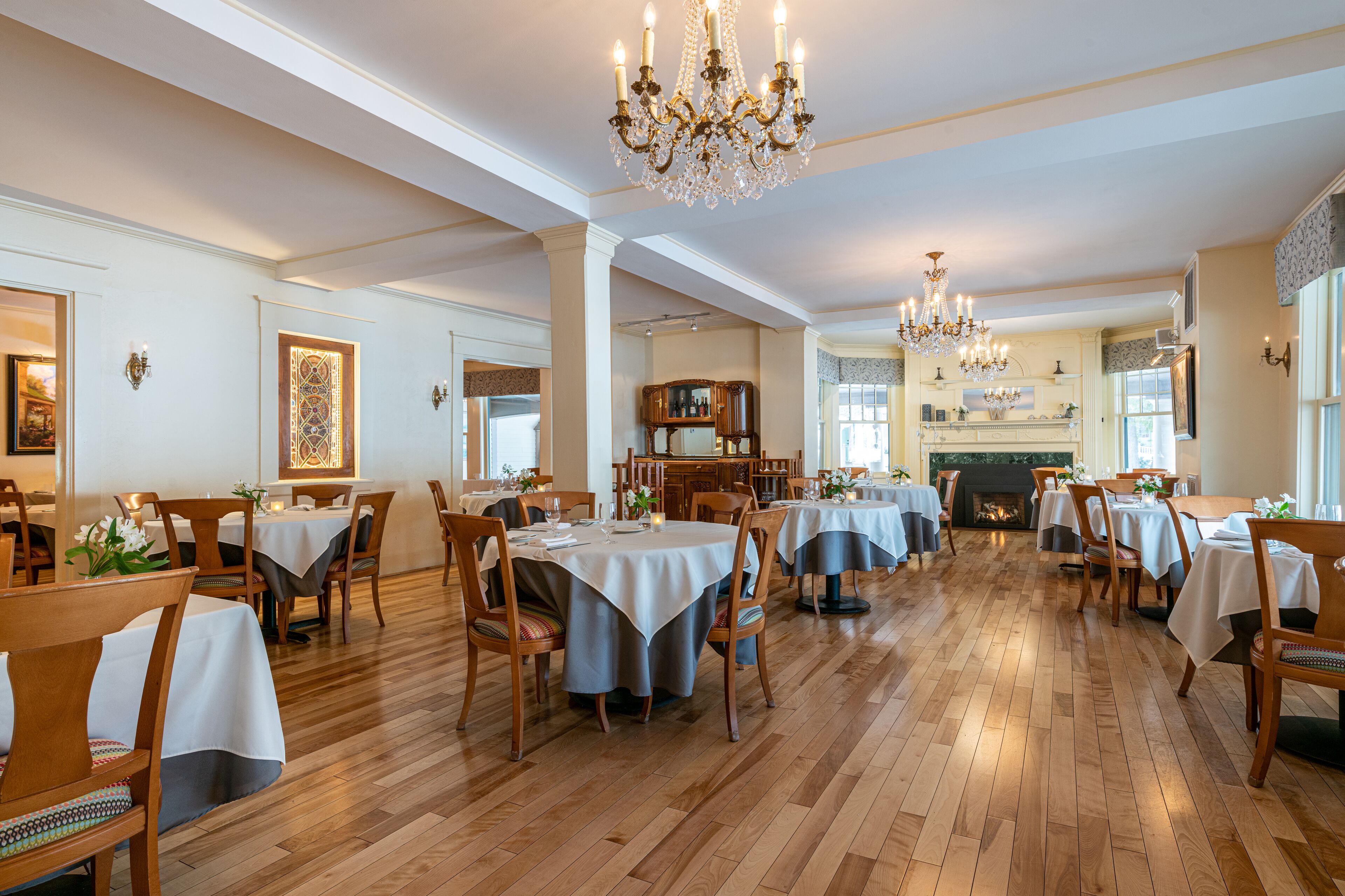 Une salle à manger spacieuse avec un plancher en bois, des tables recouvertes de nappes blanches et des lustres.