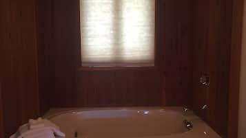 Une salle de bain aux murs lambrissés, dotée d’une grande baignoire, d’une fenêtre avec des stores et d’une pile de serviettes blanches.