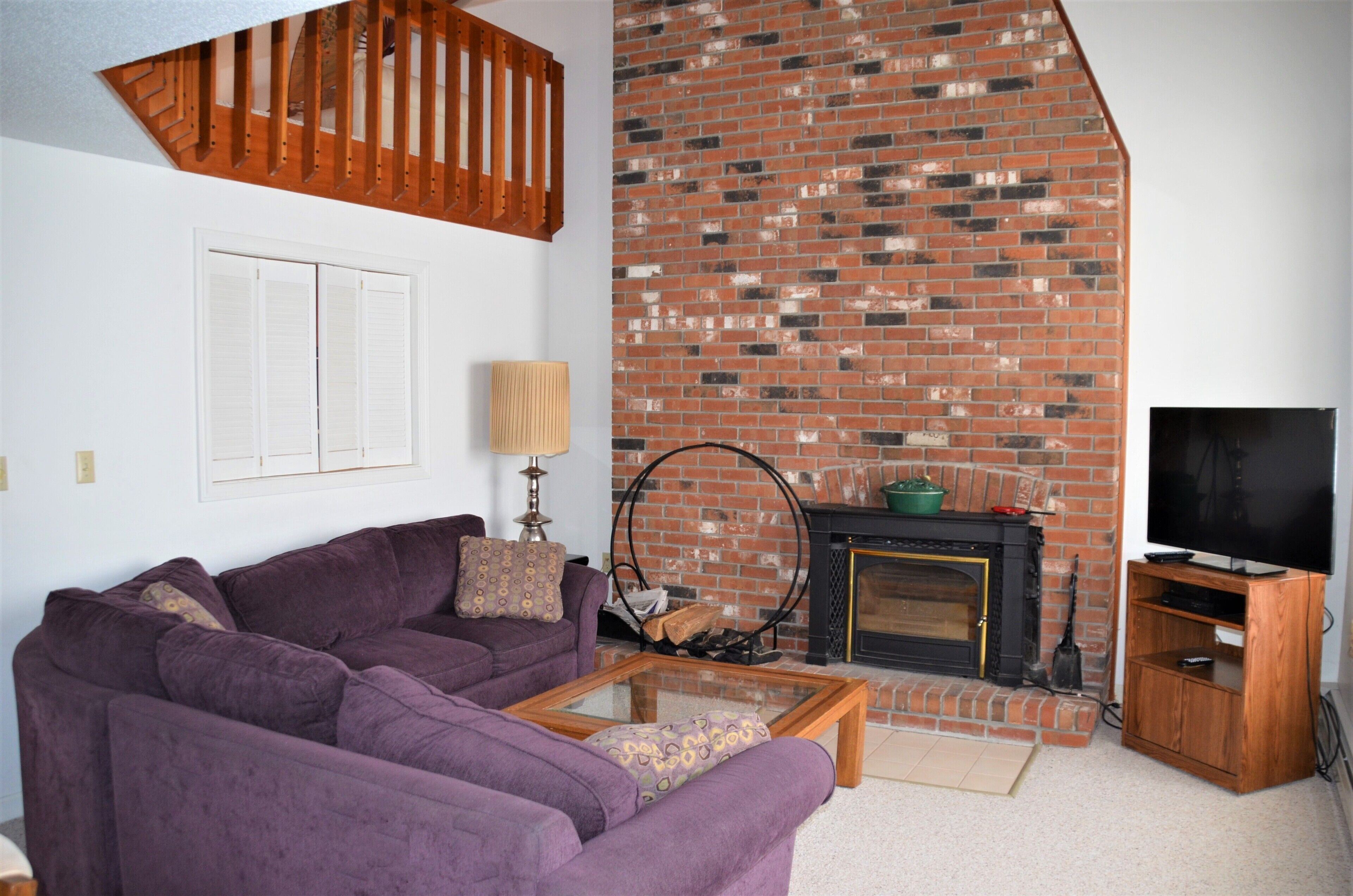 Una sala de estar con un sofá morado, una mesa de centro de vidrio, una chimenea de ladrillo y un televisor sobre un mueble de madera.
