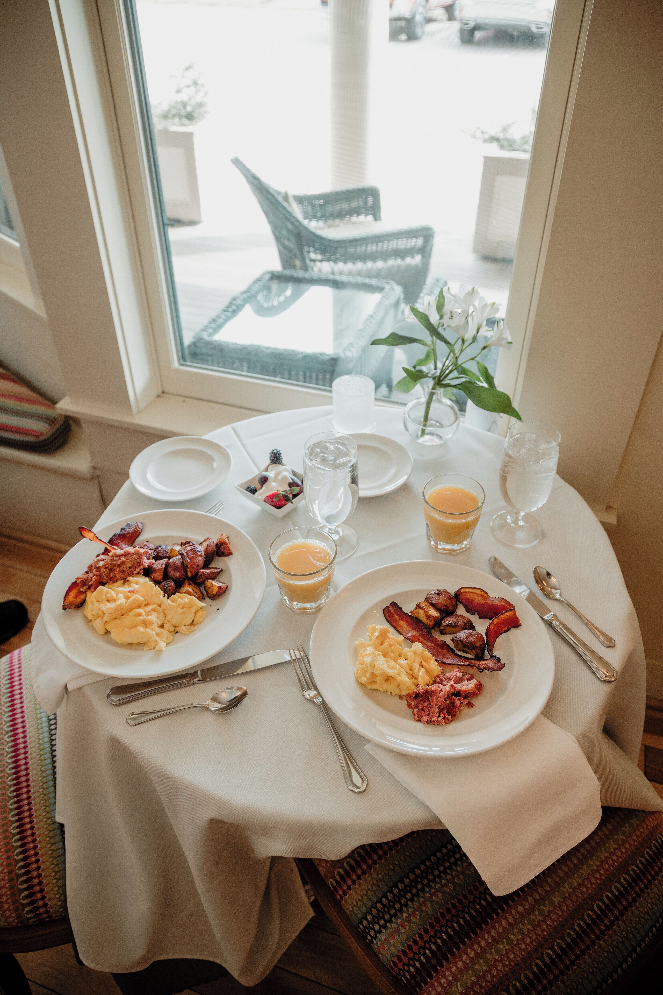 A breakfast table set with plates of eggs, bacon, and toast, along with glasses of orange juice and water, in a room with a view of the street outside.