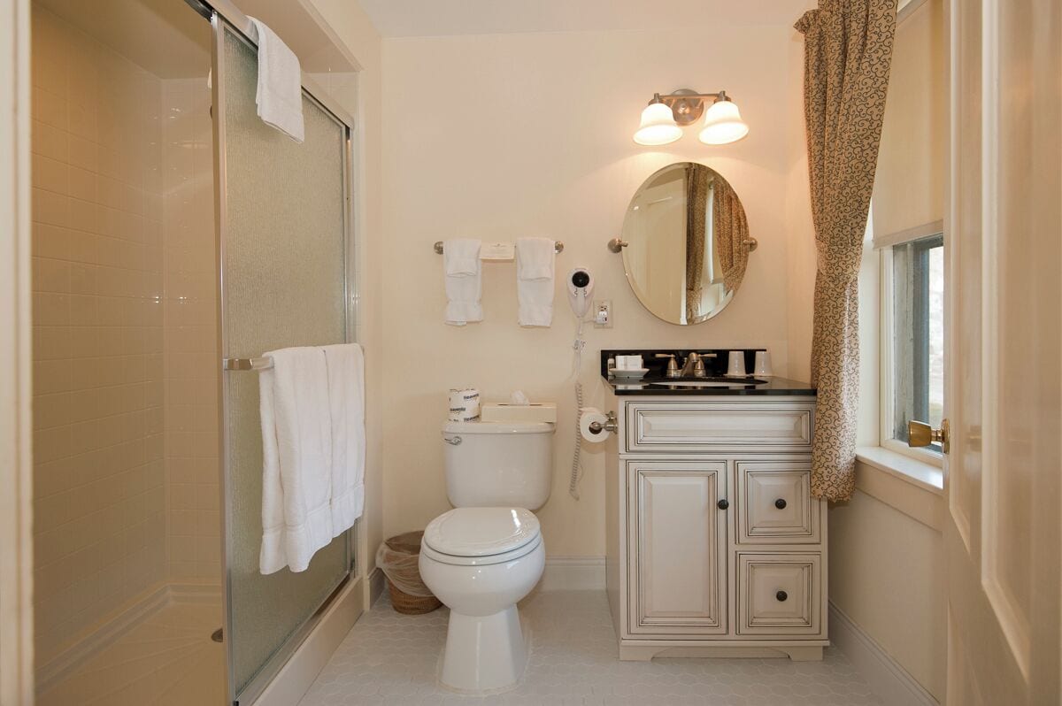 Un baño con cabina de ducha de vidrio, inodoro, tocador con espejo y ventana con cortina.