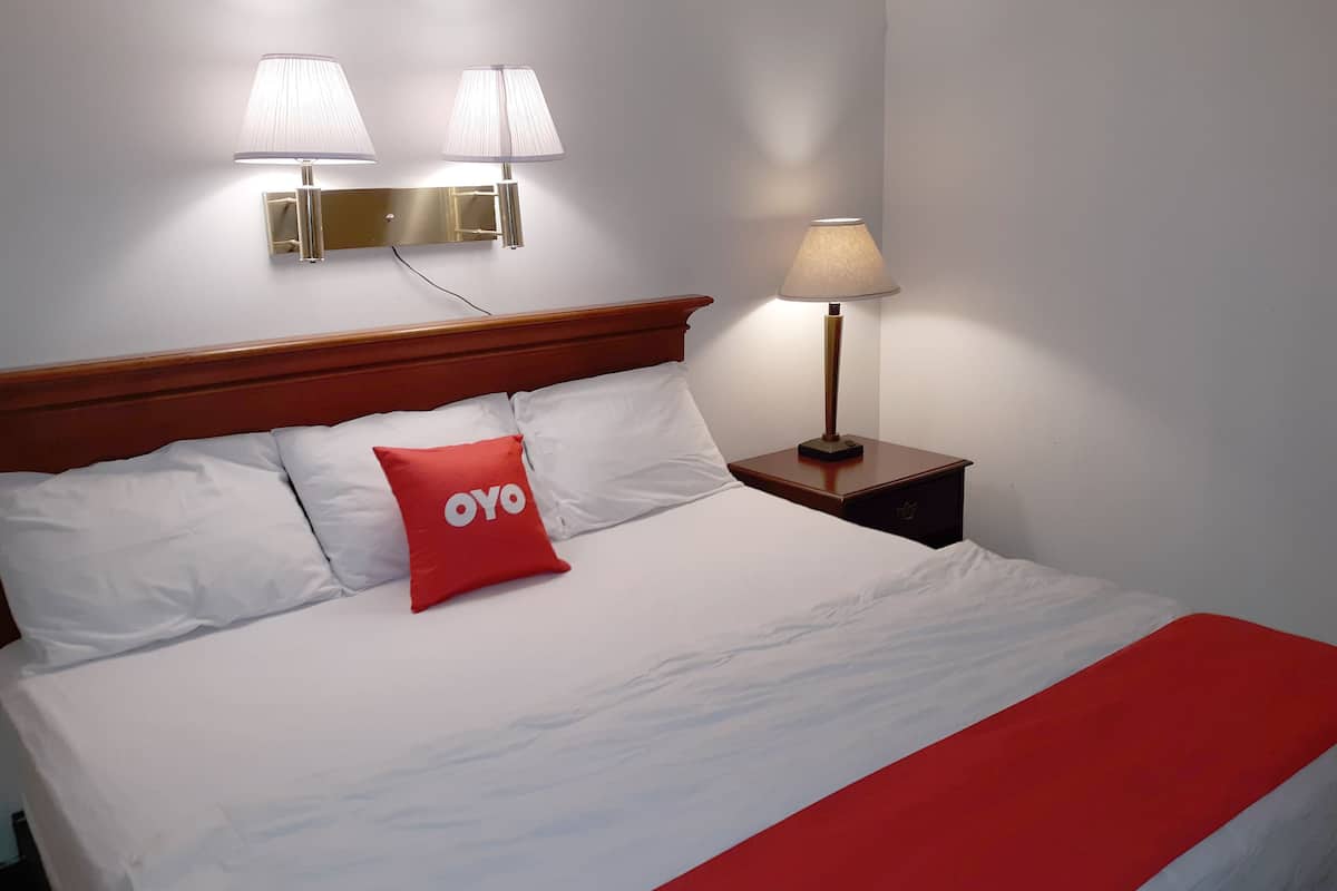 A neatly made bed with white bedding and a red pillow, a wooden headboard, and two wall-mounted lamps.