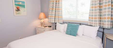 Un dormitorio con cama, una mesita de noche con lámpara, una ventana con cortinas a lunares y un cuadro con temática playera en la pared.